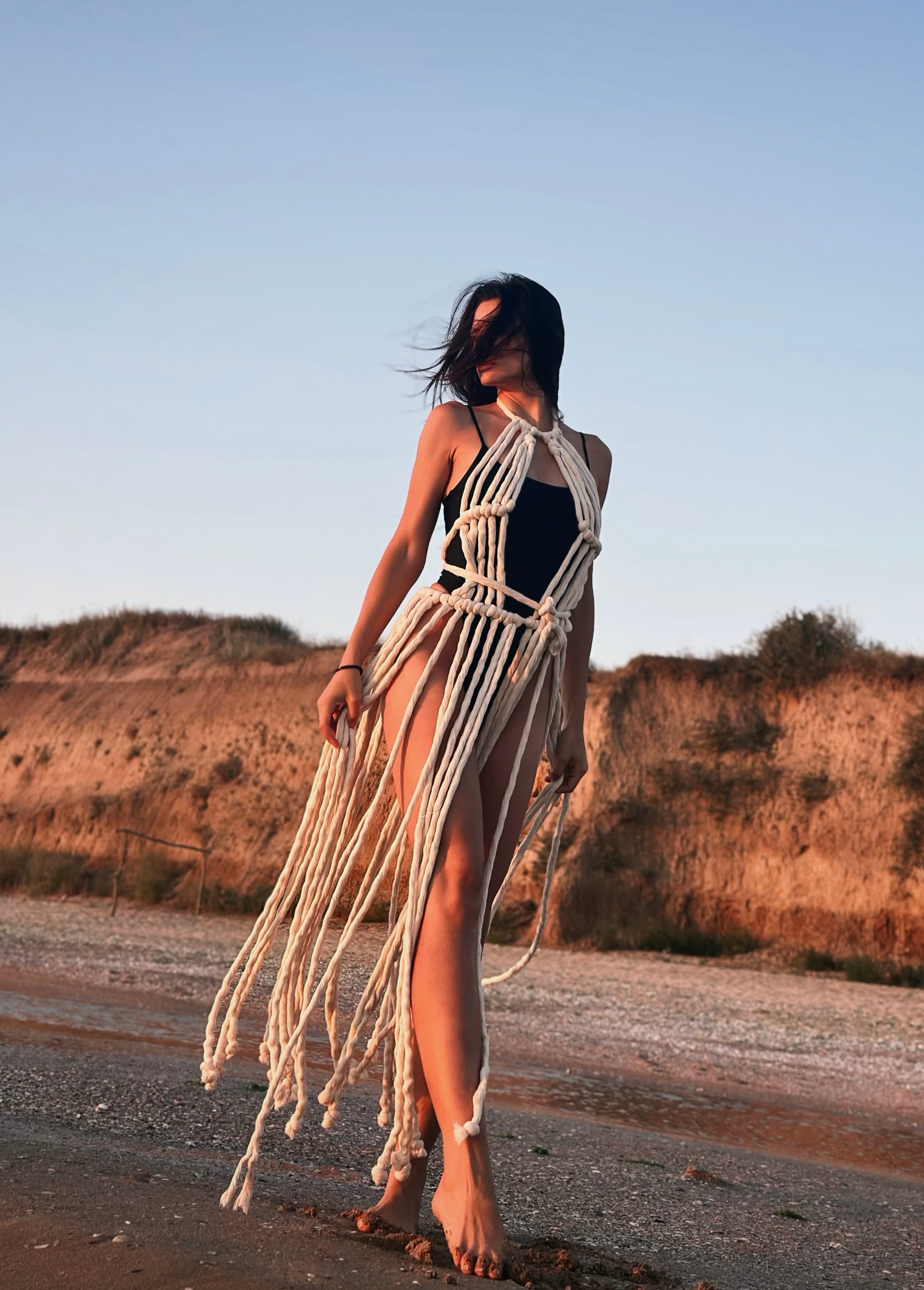 A woman stands on a sandy beach wearing a black swimsuit layered with a cream macrame fringe dress.