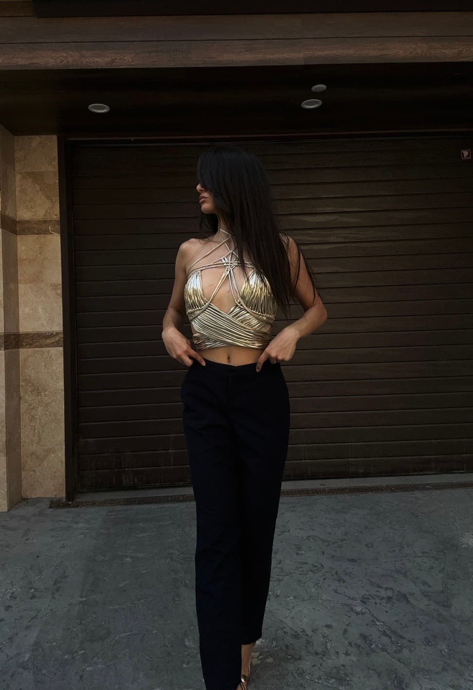 A woman wearing a shiny golden metallic bustier top with straps and black trousers outside.